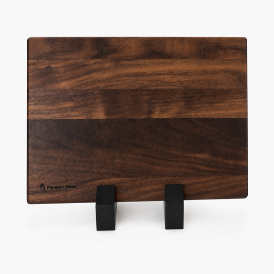 A rectangular dark wood cutting board with a prominent horizontal grain pattern, displayed on a minimalist black stand against a clean white background. The brand name 'Penguin Island' is subtly printed in black on the bottom-left corner of the board.